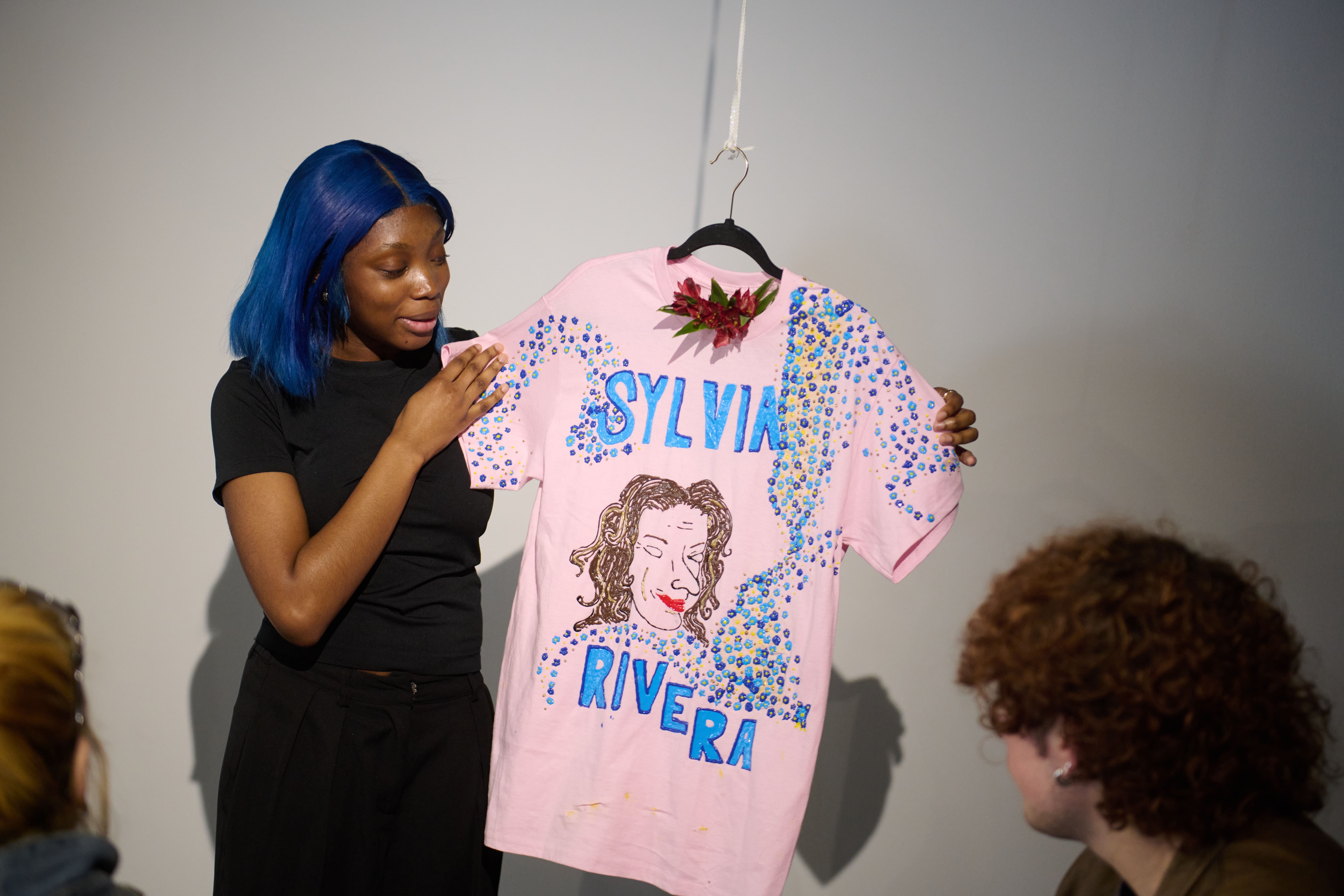 A student stands in front of seated guests and shares about the hanging artwork, a pink t-shirt decorated with puff paint in honor of Sylvia Rivera with a flower tucked into the neck.