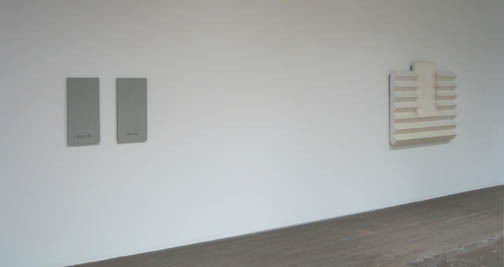 A gray painting and a cream sculptural painting stick out from a white wall.