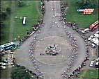 Grainy aerial view of a wide street with bikers riding past people standing on the side of the street near the grass.