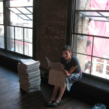 A stack of newspapers in a room with big open windows, and a person sitting down reading one.