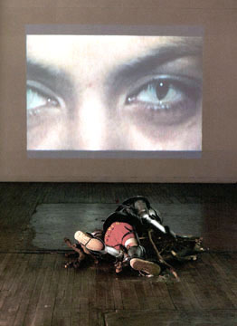 A close-up image of an eye projected on a wall, with a sculpture work on the floor below it.