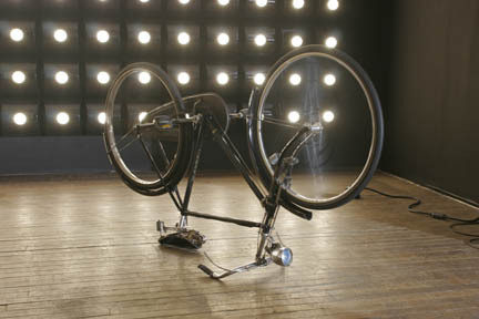 A room with lots of small lights coming from the walk, and sculptural works, bike, on the floor.