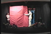An image of two people standing on a black stage moving a red curtain next to a silhouette of a female body.