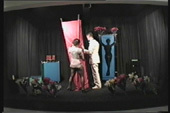 An image of two people standing on a black stage moving a red curtain next to a silhouette of a female body.