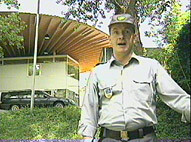 An officer, standing in front of a low-angled camera, surrounded by nature and buildings.