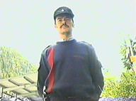 An officer, standing in front of a low-angled camera, surrounded by nature and buildings.