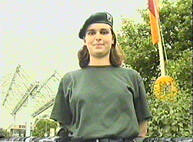 An officer, standing in front of a low-angled camera, surrounded by nature and buildings.