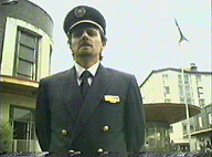 An officer, standing in front of a low-angled camera, surrounded by nature and buildings.